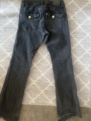 Svarta Perfect Jeans från Gina Tricot - Snygga svarta jeans från Gina Tricot, modell Perfect Jeans. Jeansen har utsvängda ben och låg midjade och  två bakfickor med lock och silverfärgade knappar. Klassisk femficksmodell i stretchigt denimtyg som sitter skönt och passar till många olika stilar.