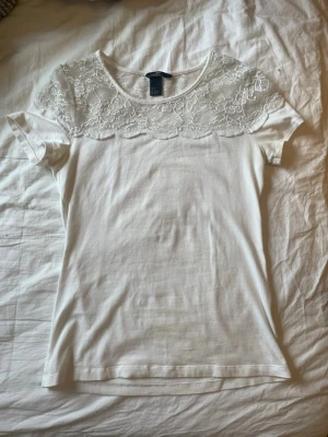 Vit topp med spets  - Säljer en vit blus från H&M med korta ärmar och snygg spets upptill över axlar och bröst. Blusen har en rund halsringning och är gjord i mjukt material. Perfekt för dig som gillar en clean look med lite extra detalj.