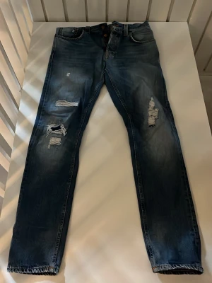 Blå slitna jeans från Nudie Jeans - Säljer ett par blå jeans från Nudie Jeans med snygga slitningar och hål på benen. Storlek 31:34. Nypris: 1399kr Mitt pris: 349