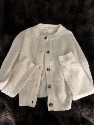 Krämvit stickad kofta med knappar - Mysig krämvit stickad kofta med rund hals och stora bruna knappar framtill. Koftan har en avslappnad passform och ribbade muddar vid ärmslut och nederkant. Perfekt för lager på lager och enkel att styla till olika outfits. Endast andvänd ett fåtal gånger!