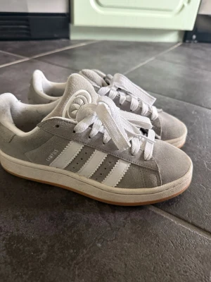 Adidas Campus grå sneakers - Säljer ett par grå Adidas Campus sneakers med klassiska vita ränder på sidan, vit sula och vita skosnören. Ovandelen är i mocka och modellen har en låg profil som ger en clean och tidlös look.