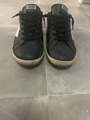 Golden Goose svart/vit sneakers - Svarta sneakers från Golden Goose med vit stjärndetalj på sidan och vit häl. Modellen heter Super-Star och har svarta skosnören samt en låg silhuett. Skorna har en klassisk design med gummisula och är tillverkade i läder.