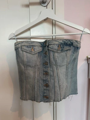 Tubtopp i denim med knappar - Säljer en snygg tubtopp i ljusblå denim med knappar framtill och rå kant nertill. Toppen har två dekorativa bröstfickor och smockad baksida för skön passform. Perfekt till sommaren och lätt att styla med olika accessoarer.