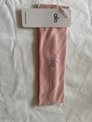 Rosa hårband från Alo Yoga - Snyggt och stretchigt rosa hårband från Alo Yoga med diskret logga framtill. Tillverkat i mjukt material som sitter bekvämt på huvudet och håller håret på plats. Perfekt accessoar för att lyfta din look och hålla håret ur ansiktet.