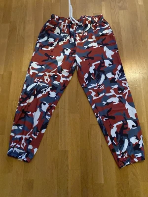 Camo-mönstrade mjukisbyxor - Säljer ett par mjukisbyxor med röd, blå, vit och svart camo-mönster. Byxorna har resår i midjan med vit dragsko och resår vid bensluten. Perfekta för en avslappnad och cool stil.