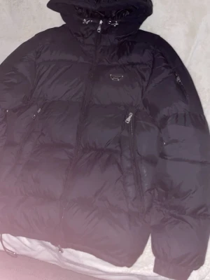 Svart pufferjacka Alessandro Zavetti - Snygg svart pufferjacka från Alessandro Zavetti med huva och flera dragkedjeförsedda fickor. Jackan har ett metallmärke på bröstet och quiltad design som ger en modern look. Perfekt för kalla dagar och riktigt varm.