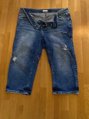 Blå jeansshorts med nitar och slitningar - Säljer ett par blå jeansshorts från Lindex med coola slitningar och nitar på fickorna. Shortsens denim är i klassisk blå färg och har en avslappnad passform. Perfekta för en avslappnad stil under varmare dagar.