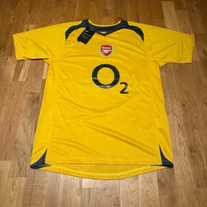 Arsenal bortatröja Henry #14 gul - Gul Arsenal bortatröja från Nike med svarta detaljer och tryck. På bröstet finns Arsenal-loggan och O2 som sponsor, på ryggen står det Henry och nummer 14. Tröjan har korta ärmar och är tillverkad i lätt polyester, perfekt för fotboll.