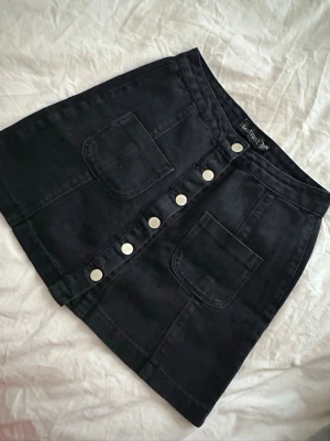 Svart kortkjol med knappar  - Snygg svart kortkjol från Isay Rose i denim med stora framfickor och silverfärgade knappar längs hela framsidan. Kjolen har en rak passform och är perfekt för en trendig och avslappnad stil.