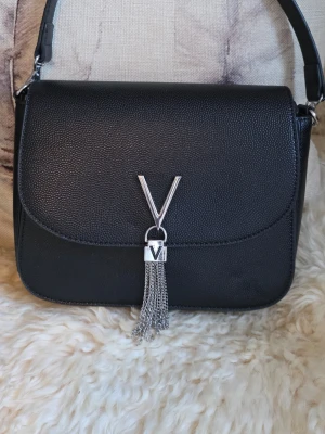 Valentino - Snygg svart handväska från Valentino med silverfärgad V-detalj och dekorativa kedjor framtill. Väskan har en stilren, fyrkantig form och är gjord i strukturerat konstläder. Perfekt accessoar för att lyfta din outfit. Väskan är i fint skick. 