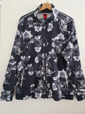 Svart Adidas blommig träningsjacka XXL - Snygg svart Adidas träningsjacka med hel dragkedja och vitt blommönster i olika nyanser. Jackan har hög krage, långa ärmar och vita detaljer längs sidorna. Perfekt för dig som vill sticka ut med en unik och trendig look. Storlek XXL. Använd, bra skick