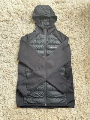 Svart hybridjacka Peak Performance - Snygg svart hybridjacka från Peak Performance med quiltad front och huva. Jackan har dragkedja hela vägen, diskret logga på bröstet och är tillverkad i polyester med vadderade partier. Perfekt för lager-på-lager och outdoor.