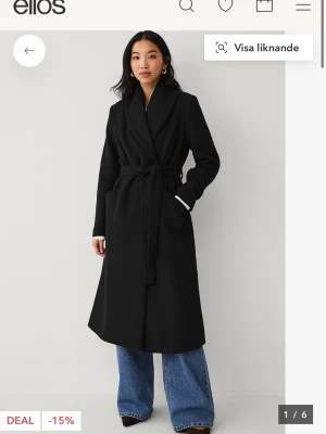 Lång kappa - Stilren svart kappa från Ellos med lång, rak passform och bred sjalkrage. Kappan har knytskärp i midjan och två sidfickor. Perfekt inför våren och enkel att matcha med olika outfits. 