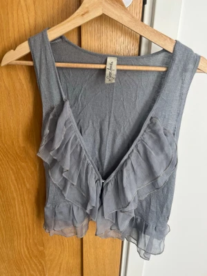 Supersöt grå volang topp från Shere Female - Supersöt grå linnetopp från Shere Female med volangdetaljer framtill. Toppen är ärmlös och har en croppad passform med transparenta volanger som ger en romantisk vibe. Perfekt att styla med höga jeans eller kjol.