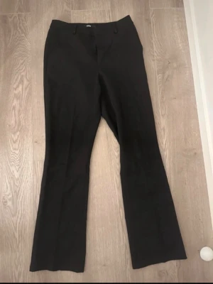 Svarta utsvängda kostymbyxor - Snygga svarta kostymbyxor med utsvängda ben och medel midja. Klassisk design med bälteshällor och diskret knappstängning framtill. Perfekta för en clean och stilren look. Materialet känns mjukt och följsamt. Passar längd runt 170.