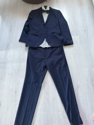 Mörkblå kostym från Jack & Jones - Snygg mörkblå kostym från Jack & Jones Premium i slim fit-modell. Kavajen har två knappar, klassiska fickor och matchande byxor. Perfekt för dig som tar studenten. Byxorna är i storlek 56 och kavajen är i storlek 56. Skjortan är en L. Bara använd en gång 