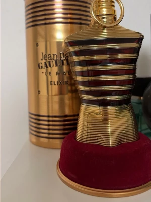 Jean Paul Gaultier Le Male Elixir - Unik parfymflaska formad som en manlig torso i guld och svart med horisontella ränder. Flaskan står på en rund röd sammetsbas och levereras i en matchande guldfärgad metalltub. En ikonisk doft för dig som gillar exklusiv design.