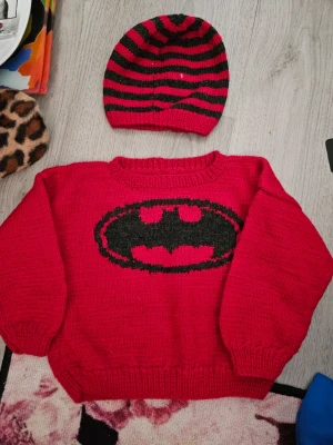 Röd Batman stickad tröja & mössa - Stickad röd tröja med svart Batman-logga på bröstet och matchande röd-svart randig mössa. Tröjan har rund halsringning, långa ärmar och ribbstickade muddar. Perfekt för dig som gillar superhjältar och vill ha en unik look.