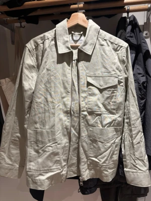 Beige overshirt med fickor - Stilren beige overshirt med dragkedja framtill och tre stora fickor, varav en bröstficka med lock. Overshirten har klassisk krage och raka ärmar. Perfekt för lager-på-lager och enkel att matcha med olika outfits. Materialet ser ut att vara bomull. Behövs strykas/steamas.