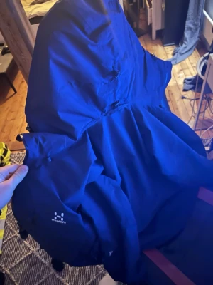 Blå vindjacka från Haglöfs - Snygg blå vindjacka från Haglöfs med minimalistisk design och justerbar huva. Jackan har diskret logga på sidan och är tillverkad i ett lätt, vindtätt material som passar perfekt för utomhusaktiviteter. Praktisk och stilren modell.