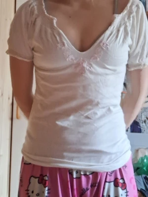 Gräddvit T-shirt med blommigt detalj  - Säljer en bekväm T-shirt i en gräddvit nyans med ljusrosa blom-detaljer längs en intressant ringning. Säljer då den aldrig kommer till användning, aldrig använd men eventuellt begagnad. Vill bli av med allt så kan sälja billigt. Köpare föreslår pris 🍀
