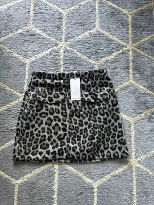 Leopardmönstrad kortkjol från cherry koko - Snygg kortkjol från & cherry koko med leopardmönster i svart och beige. Kjolen har två dekorativa fickor med knappar framtill och är gjord i ett mjukt material som känns lite ulligt. Perfekt statement-plagg för dig som vill sticka ut.