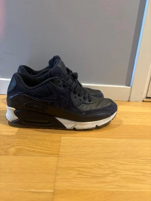 Blå Nike Air Max 90 sneakers - Säljer ett par mörk blå Nike Air Max 90 sneakers med klassisk chunky sula och synlig Air-enhet. Skorna har en mix av läder och mesh i ovandelen, svarta skosnören och detaljer i blå och vitt på sulan. Perfekta för dig som gillar streetwear och sportig stil.