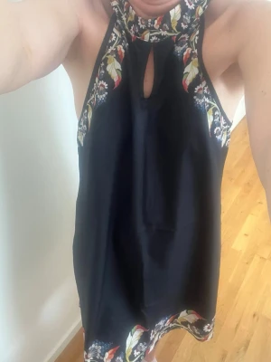 Svart blommig ärmlös kortklänning - Snygg svart kortklänning med blommigt mönster längs kanterna och halsringningen. Klänningen är ärmlös med en liten öppning framtill vid halsen och har en rak passform. Perfekt för sommarens alla tillfällen.