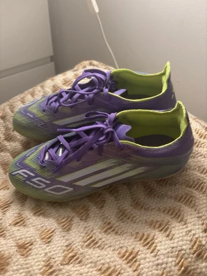 Adidas F50 lila/gröna fotbollsskor - Säljer ett par Adidas F50 fotbollsskor i en riktigt snygg lila och grön färgkombination med vita detaljer och klassiska tre ränder på sidan. Skorna har snörning och är tillverkade i ett lätt syntetmaterial som ger bra känsla på planen.