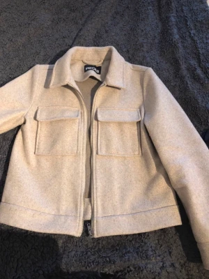 Beige kappa från Pieces - Snygg beige kappa från Pieces med dragkedja framtill och två stora bröstfickor med lock. Overshirten har klassisk krage och är tillverkad i ett mjukt, filtliknande material. Perfekt att slänga över en t-shirt för en trendig look.