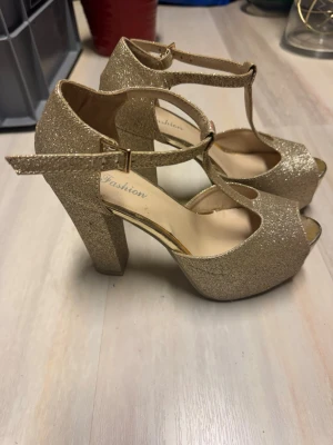 Glittriga guldiga T-rem pumps - Säljer ett par glittriga guldiga pumps med T-rem och öppen tå. Skorna har en hög platå och bred klack för extra stabilitet. Remmen runt ankeln har ett spänne i guld. Perfekta för dig som vill sticka ut och glänsa lite extra.