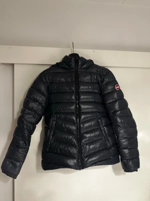 Marinblå pufferjacka med huva - Snygg marinblå pufferjacka med huva och dragkedja framtill. Jackan har två sidofickor med dragkedja och ett diskret märke på ärmen. Den quiltade designen ger en cool look och håller dig varm under kalla dagar. Perfekt för dig som vill ha en stilren och praktisk vinterjacka/höstjacka