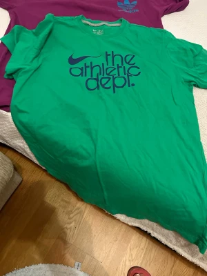 Grön Nike t-shirt Athletic Dept - Säljer en grön t-shirt från Nike med trycket 'the athletic dept.' i mörkblått på bröstet. T-shirten har klassisk rund hals och korta ärmar, tillverkad i mjuk bomull. Perfekt för dig som gillar sportiga och avslappnade looks.