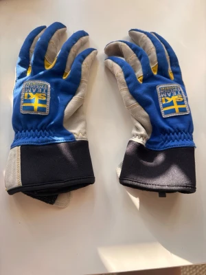 Hestra vantar - Fina längdskids handlar med lite användar märken på finger topparna. Snygga blå och grå handskar från Team Sweden med svensk flagga och Wind Stopper-detaljer. Handskarna har kardborreband vid handleden för bra passform och är tillverkade i slitstarkt skinn och syntetmaterial. Perfekta för dig som vill ha både stil och funktion.