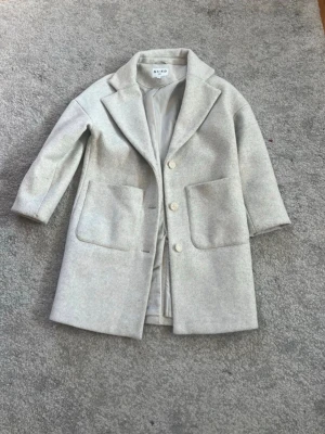 Ljusgrå kappa från NA-KD - Stilren ljusgrå kappa från NA-KD med stora framfickor och klassisk krage. Kappan har två knappar framtill och är fodrad på insidan. Perfekt för dig som gillar minimalistisk och trendig stil.