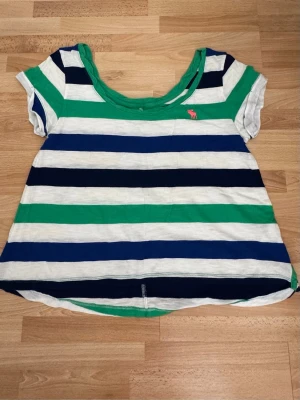 Randig topp från Abercrombie & Fitch - Kortärmad topp med breda gröna, vita och marinblå ränder från Abercrombie & Fitch. Rund halsringning och avslappnad passform. Liten rosa logga broderad på bröstet. Perfekt för en chill och casual stil. Använd en gång. 
