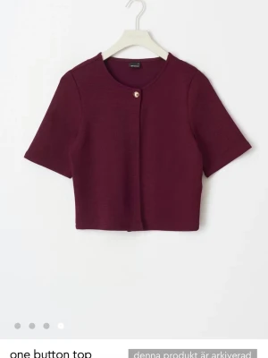 Vinröd croppad topp med knapp - Trendig vinröd croppad topp med korta ärmar och en snygg knapp framtill. Perfekt att styla över ett linne eller t-shirt under. Säljer den pga att jag knappt andvänder den men tycker att den är jätte snygg. Den är i jätte bra skicka då jag har bara andvändigt den 3 gånger totalt.