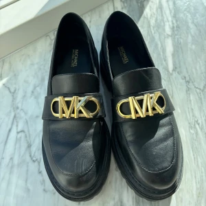 Svarta loafers med gulddetaljer från Michael Kors - Snygga svarta loafers i skinn från Michael Kors med en iögonfallande guldfärgad logga på ovansidan. Skorna har en robust sula som ger extra höjd och stil. Perfekta för att ge en elegant touch till din outfit. Ny pris är 2095 säljer för halva pris 