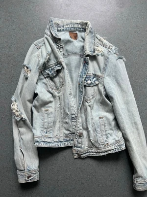 Ljusblå distressed jeansjacka AE - Säljer en ljusblå jeansjacka från American Eagle med slitna detaljer och råa kanter. Klassisk modell med bröstfickor, knappstängning och cool vintagekänsla. Perfekt för dig som gillar en avslappnad och trendig look.