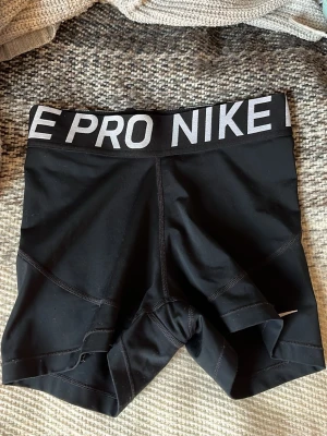 Svarta Nike Pro shorts med bred resår - Snygga svarta Nike Pro shorts med bred elastisk midjeresår där det står 'NIKE PRO' i vitt. Tight passform och tillverkade i ett stretchigt syntetmaterial som passar perfekt för träning. Klassisk sportig look och bekväma sömmar.