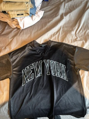 Svart oversized t-shirt New York H&M - Snygg svart oversized t-shirt från H&M med stor vit text 'NEW YORK' på bröstet. T-shirten har v-ringning och korta ärmar. Tillverkad i ett lätt och luftigt polyestermaterial som ger en sportig vibe.