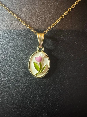 Guldhalsband med blomma och pärla - Sött halsband med tunn guldfärgad kedja och ett ovalt hänge. Hänget har en rosa blomma med gröna blad och en liten vit pärldetalj. Perfekt för dig som gillar unika och färgglada accessoarer med blommotiv.