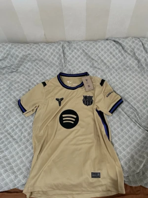 Beige FC Barcelona matchtröja - Snygg beige FC Barcelona matchtröja med svarta och blå detaljer. Tröjan har klubbmärke, sponsorlogga och Nike-logga på bröstet. Tillverkad i lätt och ventilerande funktionsmaterial, perfekt för träning eller match.