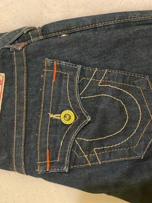 True Religion blå jeans med kontrastsömmar - Säljer ett par klassiska blå jeans från True Religion med tydliga kontrastsömmar i gult och orange. Jeansen har ikonisk fickdesign med gul knapp och detaljerad brodyr. Tillverkade i USA av slitstarkt denimtyg. Perfekta för dig som gillar streetwear och snygga detaljer.