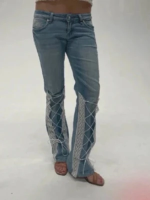 Blå bootcut jeans med spetsdetaljer - Unika blå bootcut jeans med ljus tvätt och breda spetsinslag längs benen. Snygga snörningsdetaljer framtill ger en cool och trendig look. Perfekta för dig som vill sticka ut och addera något extra till outfiten.