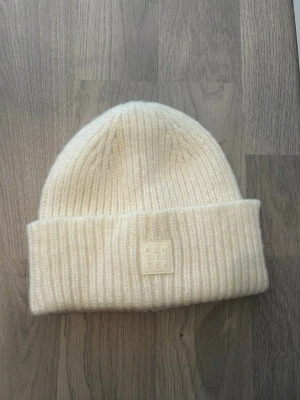 Vit ribbstickad mössa från Femi Stories - Säljer en stilren vit ribbstickad mössa från Femi Stories. Mössan har uppvikt kant och en diskret patch med logga framtill. Perfekt för kalla dagar och enkel att matcha med olika outfits. Materialet känns mjukt och värmande.
