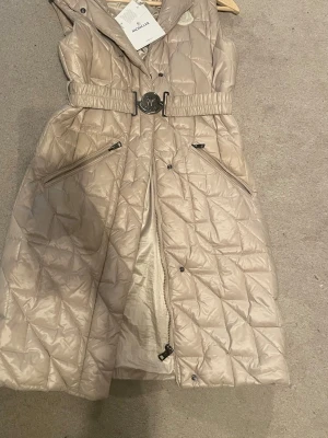 Beige lång dunväst från Moncler S - Stilren beige lång dunväst från Moncler med quiltat mönster, avtagbar huva och markerad midja med bälte. Två dragkedjefickor framtill och snyggt Moncler-emblem på bröstet. Perfekt för lager-på-lager och trendig look. Storlek: S, denna har används förra vintern / vår men är i nyskick.      Inköpt London Harvey nicols för 2 år sedan. Tags medföljer. Passa på,  Lågt utrops pris! 