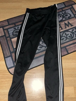 Svarta träningsbyxor med vita ränder - Säljer ett par svarta träningsbyxor med två vita ränder längs sidorna. Byxorna har resår i midjan och är tillverkade i ett lätt och stretchigt material som passar perfekt för sport och träning.