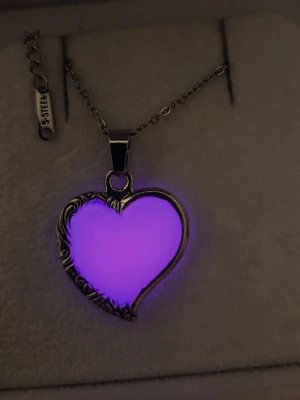 Halsband med rosa hjärthänge - Unikt halsband i rostfritt stål med ett hjärtformat hänge i starkt rosa färg som lyser lila i mörker. Hänget har dekorativa detaljer i silverfärg runt kanten och kedjan är tunn och stilren. Perfekt för dig som vill sticka ut med en cool accessoar. Vanligt dagsljus räcker för att hänget ska ändra färg och börja lysa/ge ett lila sken i mörker och lyser vanligtvis i flera timmar.