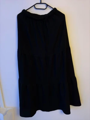 Svart långkjol med volang - En svart långkjol med volangkant och resår i midjan. Kjolen har en enkel och stilren design som passar till många olika looks. Perfekt för dig som gillar en klassisk och avslappnad stil.